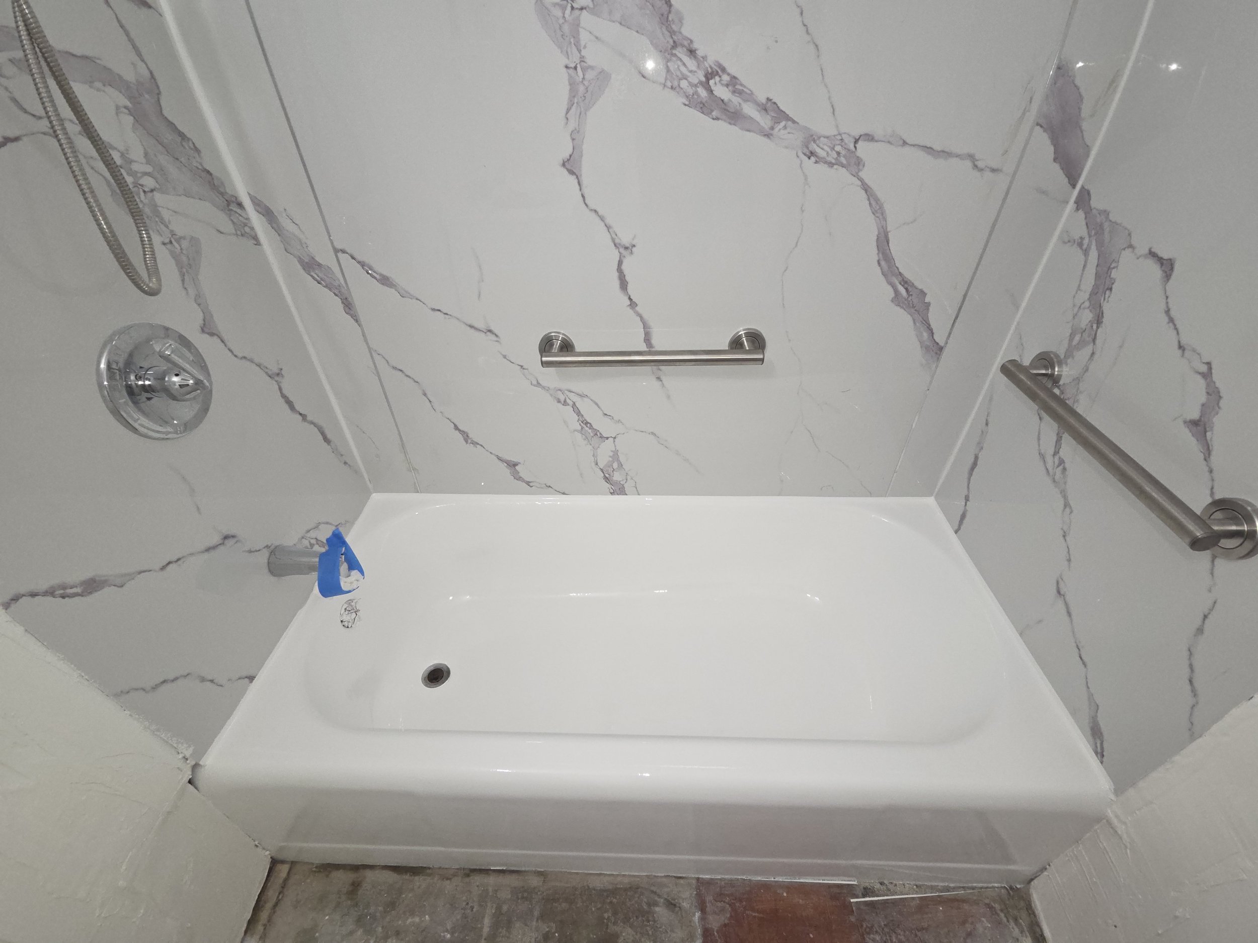 Shower and bathtub area with marble-look tile walls, a chrome showerhead, a grab bar, and a partial view of the tub with a soap holder and a toy on the edge.