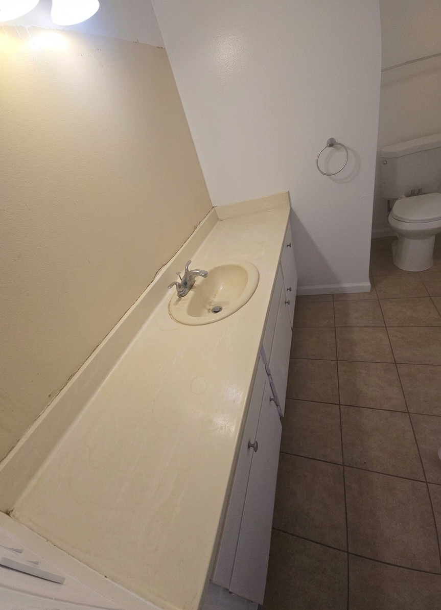 An old bathroom sink with a damaged countertop, a faucet, white cabinetry, a towel ring, and a toilet in the background.