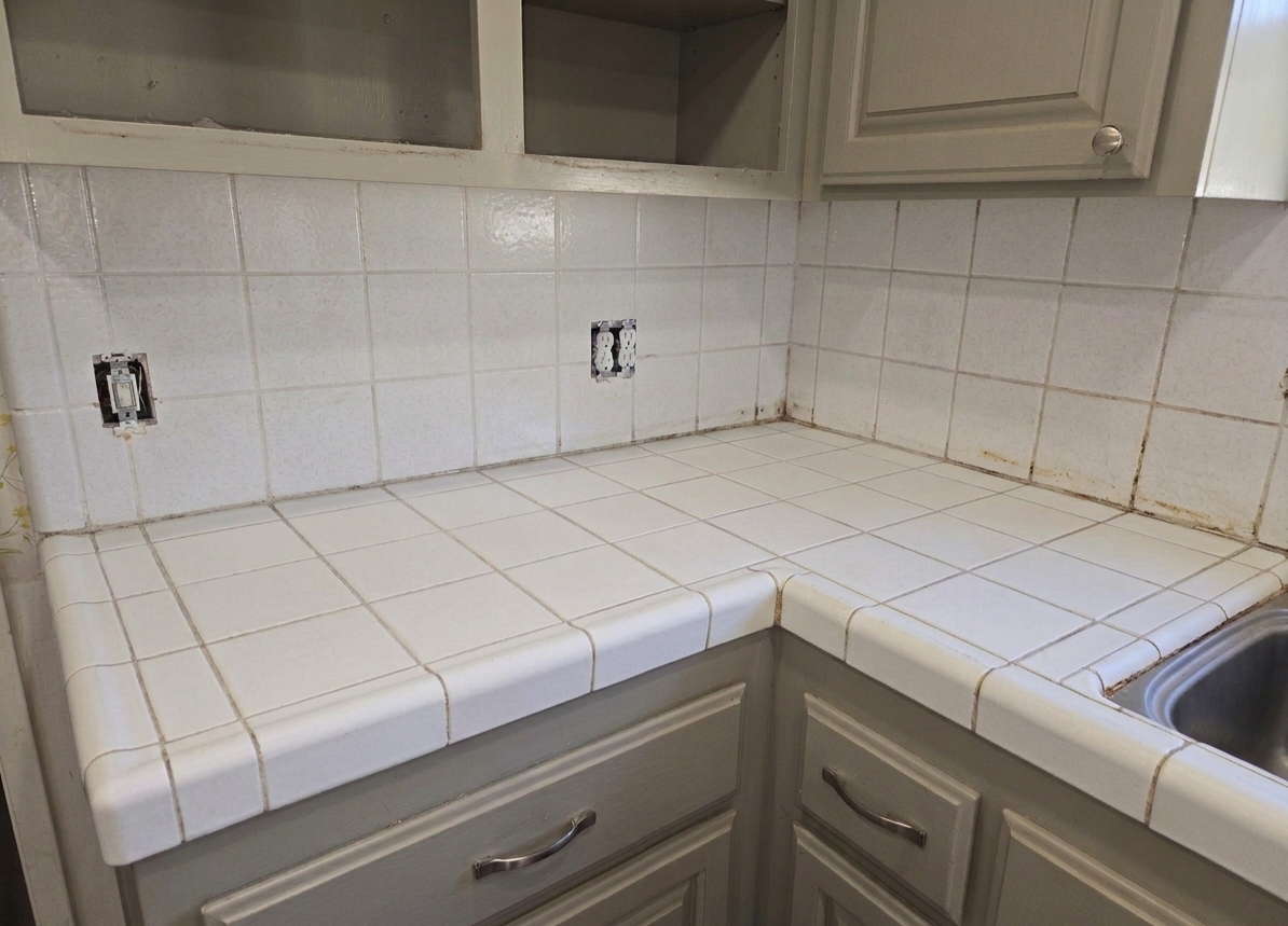 A kitchen countertop with white tiles, some grout lines are dirty, and an empty wall with electrical outlets.