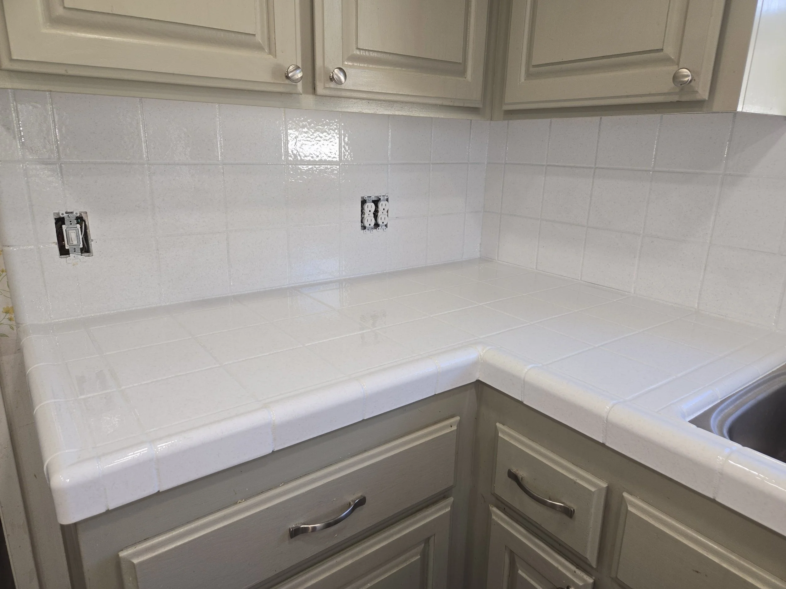 Kitchen countertop with white tiles, electrical outlets, and cabinet doors.