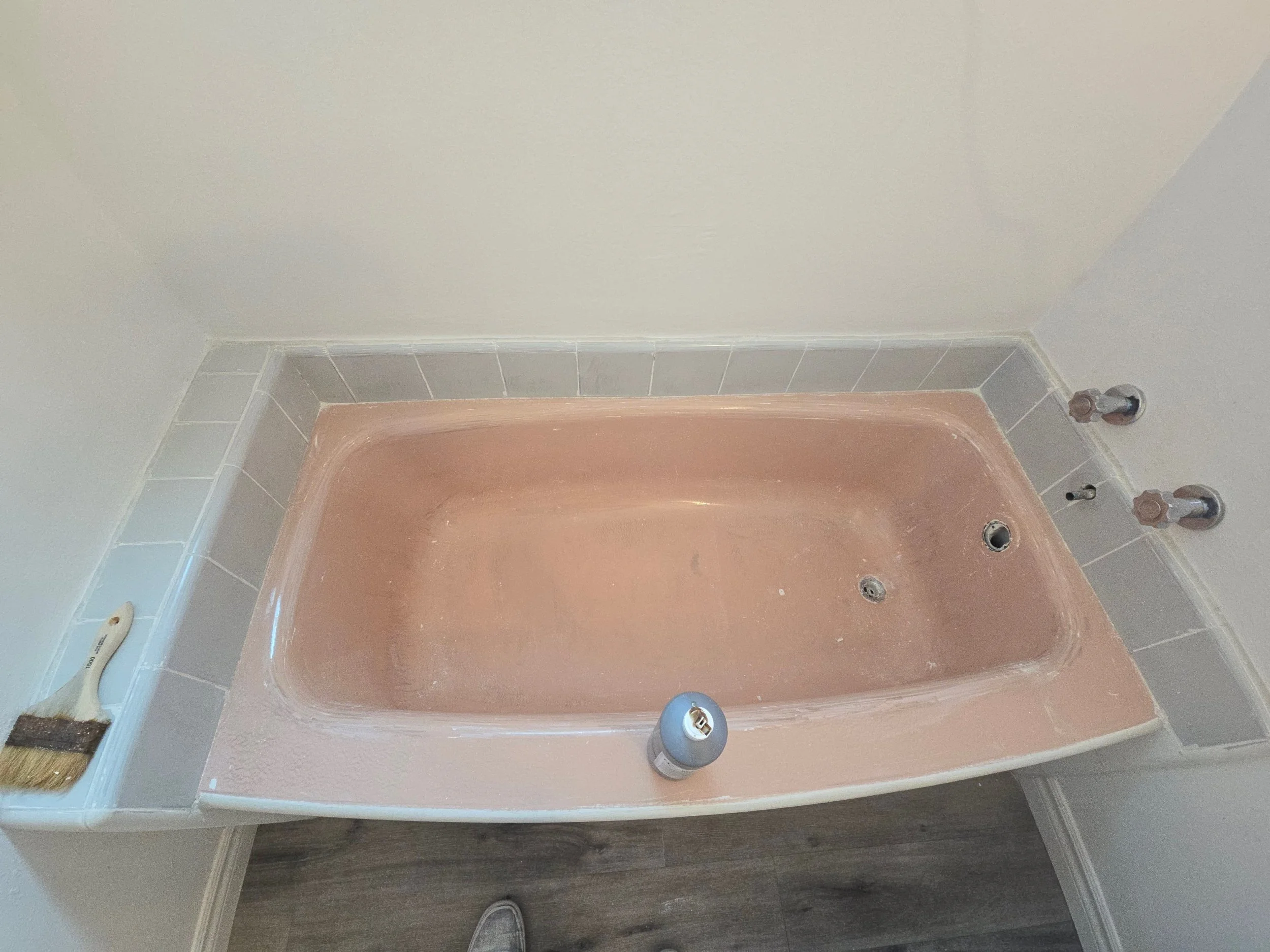Empty bathtub with a pinkish hue in a bathroom with light-colored tiled walls and a dark wood floor.