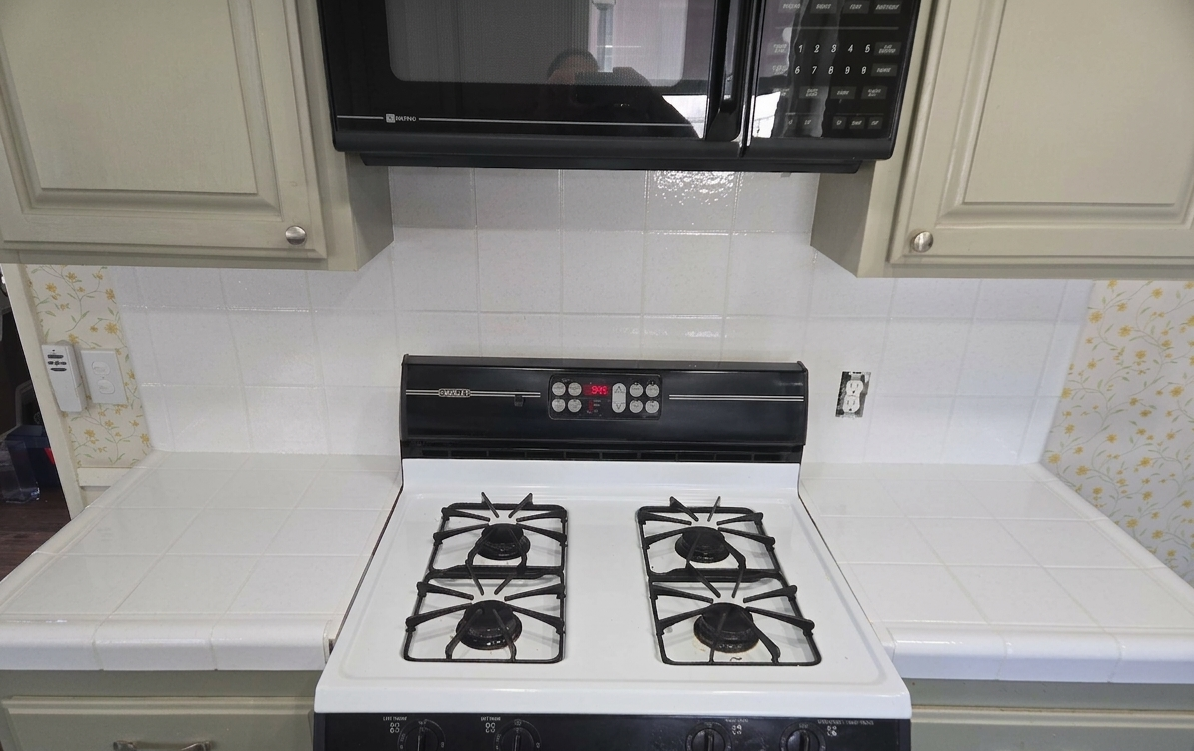 A white gas stove with four burners and black grates, situated in a kitchen with white tiled backsplash and beige cabinets above.