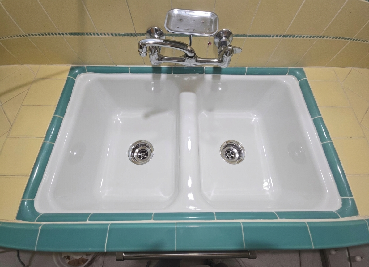 White double sink with green tile border, installed in a tiled yellow wall, featuring two drain openings and a chrome faucet above.