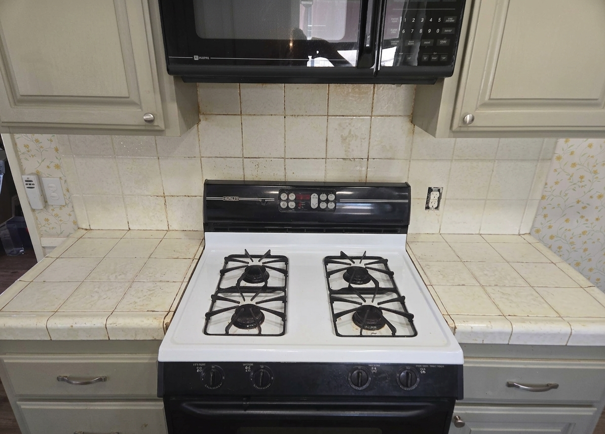 A kitchen stove with four gas burners, a black oven control panel, and a microwave above it. The surrounding countertop and backsplash have beige tiled surfaces with some dirt and staining, and a wall outlet is visible.