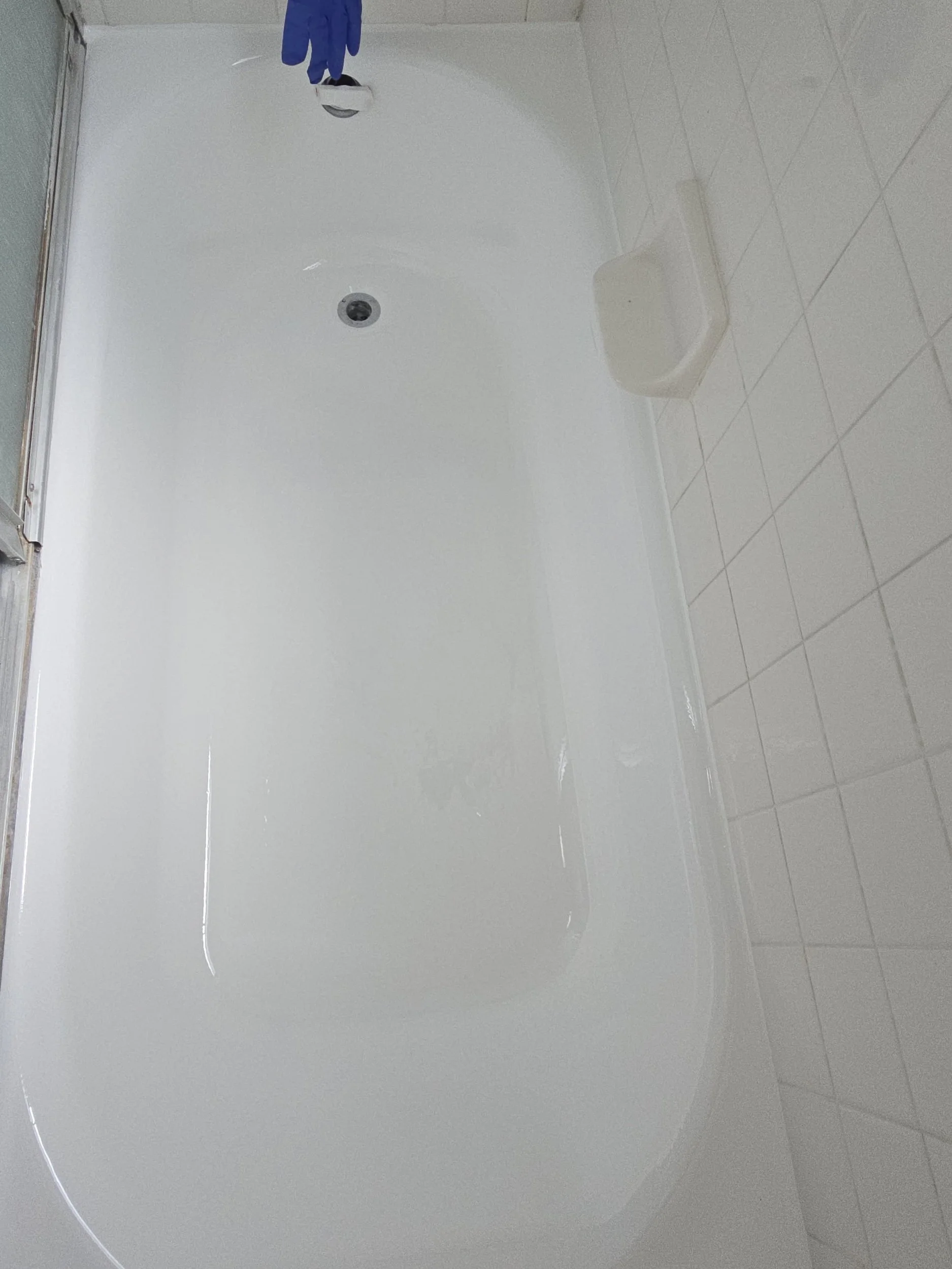 Empty shower stall with white tiled walls and a white base, with a showerhead and a pair of blue gloves hanging from the showerpipe.