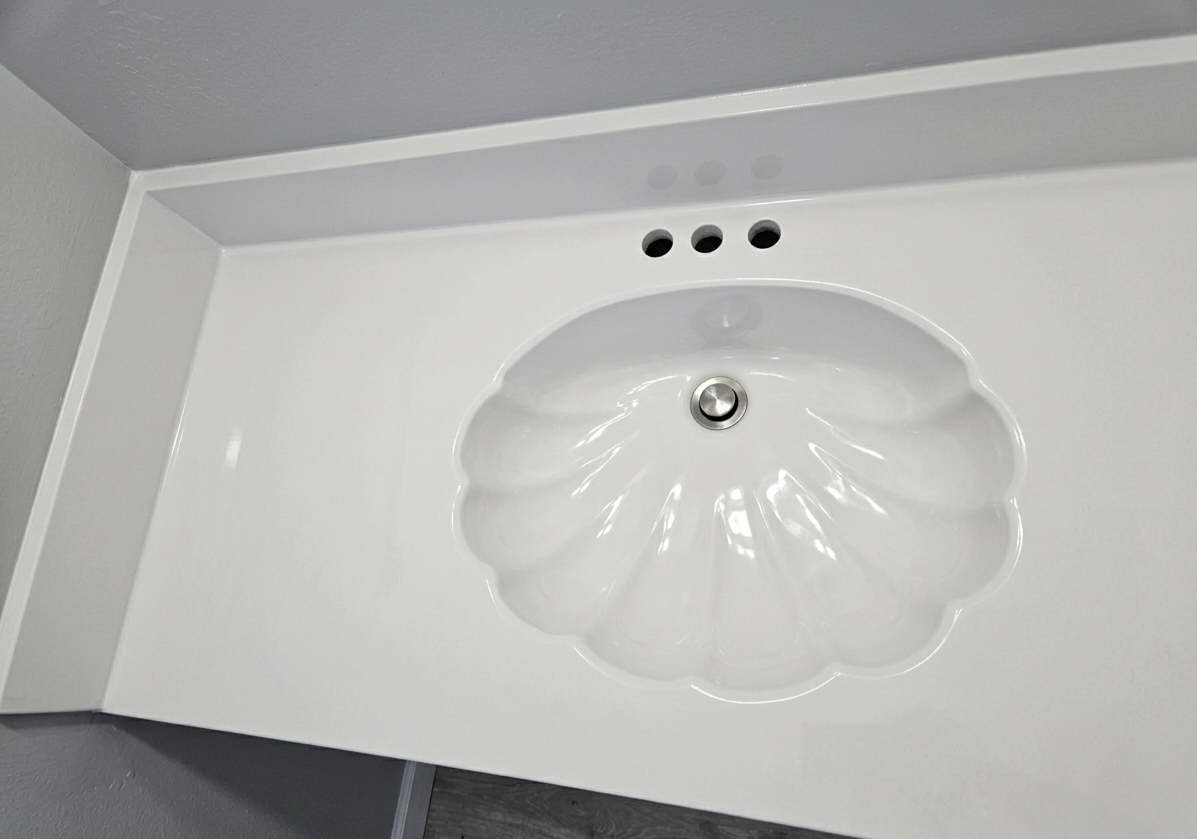 White bathroom sink with a scallop-shaped basin and three holes for faucet handles, mounted on a white counter with a built-in mirror above.