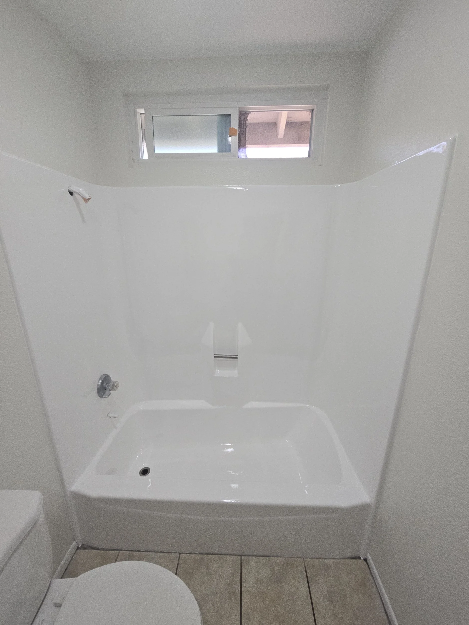 A small bathroom with a white bathtub, a white toilet, and a small horizontal window above the bathtub. The window has frosted glass on the left side and clear glass on the right side.