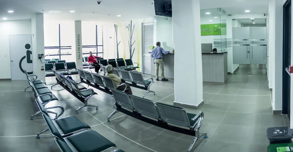Sala de espera en un hospital con sillas metálicas, pocas personas, mostrador de recepción y señalización en español.