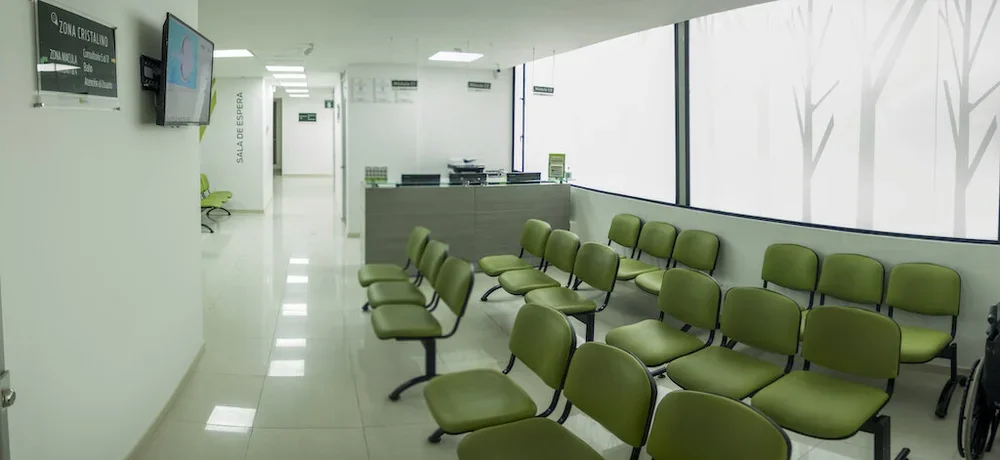 Sala de espera con sillas verdes y ventanas grandes, un mostrador y un televisor en la pared.