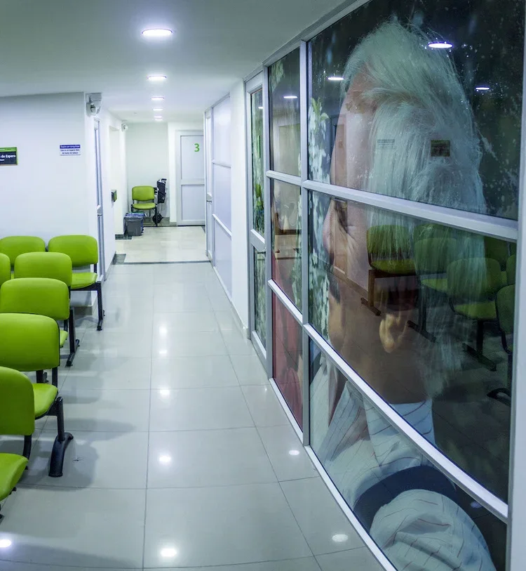 Pasillo de clínica con sillas verdes y una pared de vidrio con imagen de una mujer de cabello gris y rubio.