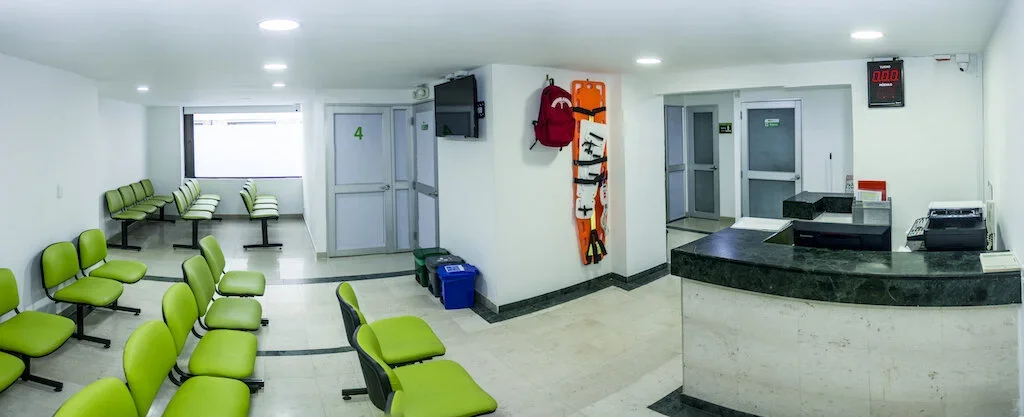 Sala de espera en una clínica o consultorio, con sillas verdes, una ventana con cortinas, un mostrador de recepción con equipo de oficina, y paredes blancas con accesorios médicos y un televisor.
