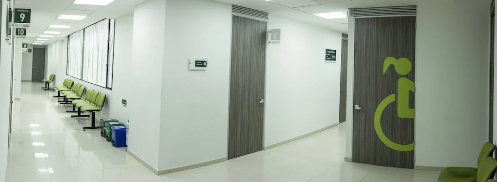 Sala de espera en un hospital con sillas verdes y una señal de acceso para discapacitados en la puerta.