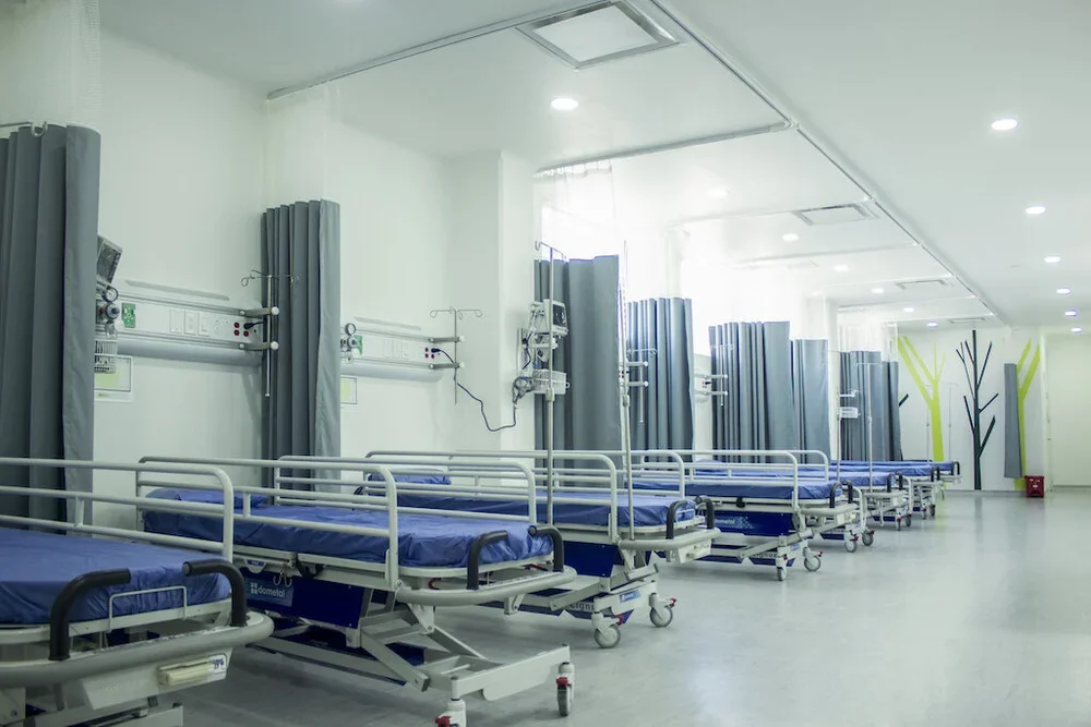 Habitaciones de hospital vacías con camas y cortinas en corredor amplio y bien iluminado.