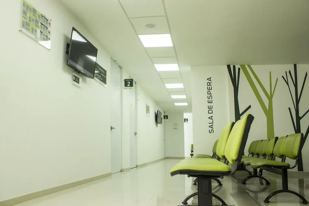 Sala de espera en un hospital con sillas verdes, paredes blancas y monitores en la pared, con un mural de árboles y la frase 'Sala de Espera'