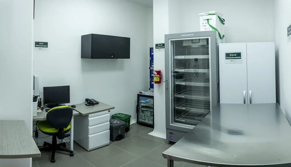 Una oficina en una área de almacenamiento y refrigeración, con un computadora, una silla, un refrigerador grande y un gabinetes blancos con señalización en español.