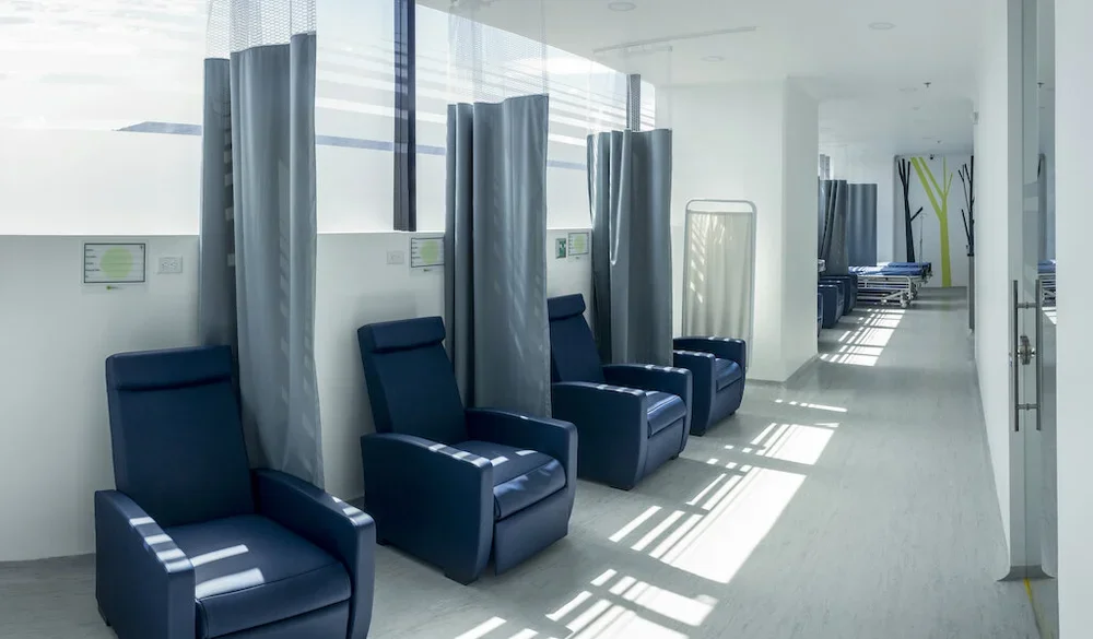 Sala de espera en un hospital o clínica con sillas azules y cortinas grises, con ventanas que dejan entrar luz natural.