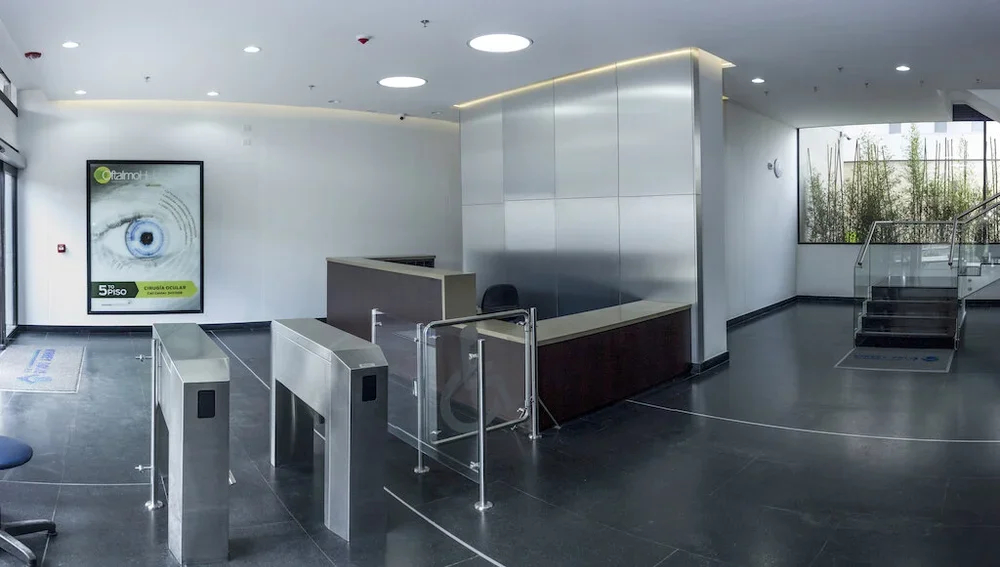 Recepción moderna en una clínica, con mostradores, puertas de acceso controlado y escalera con barandales de vidrio.