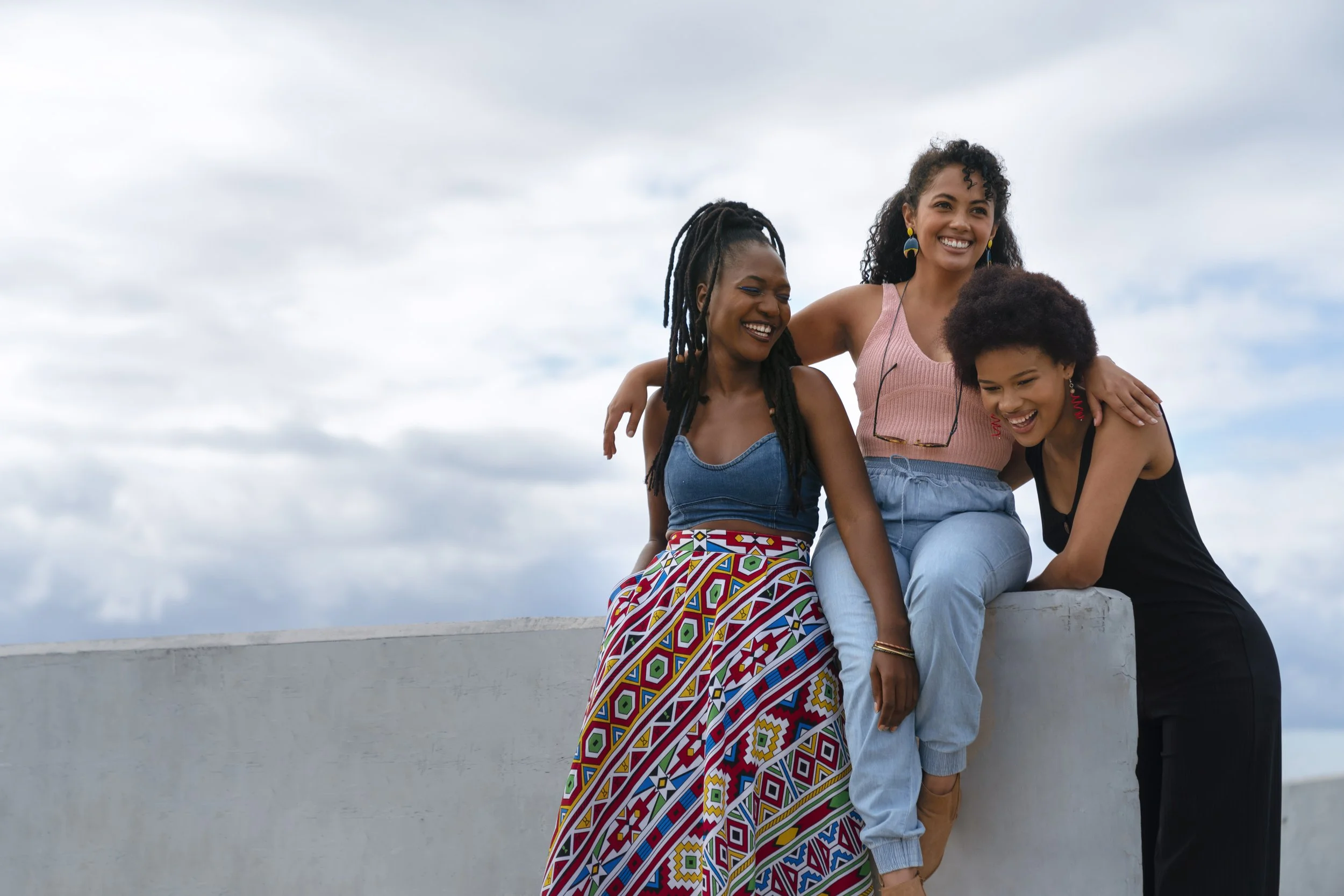 Tres mujeres sonrisas, abrazándose y disfrutando en un día con cielo nublado.