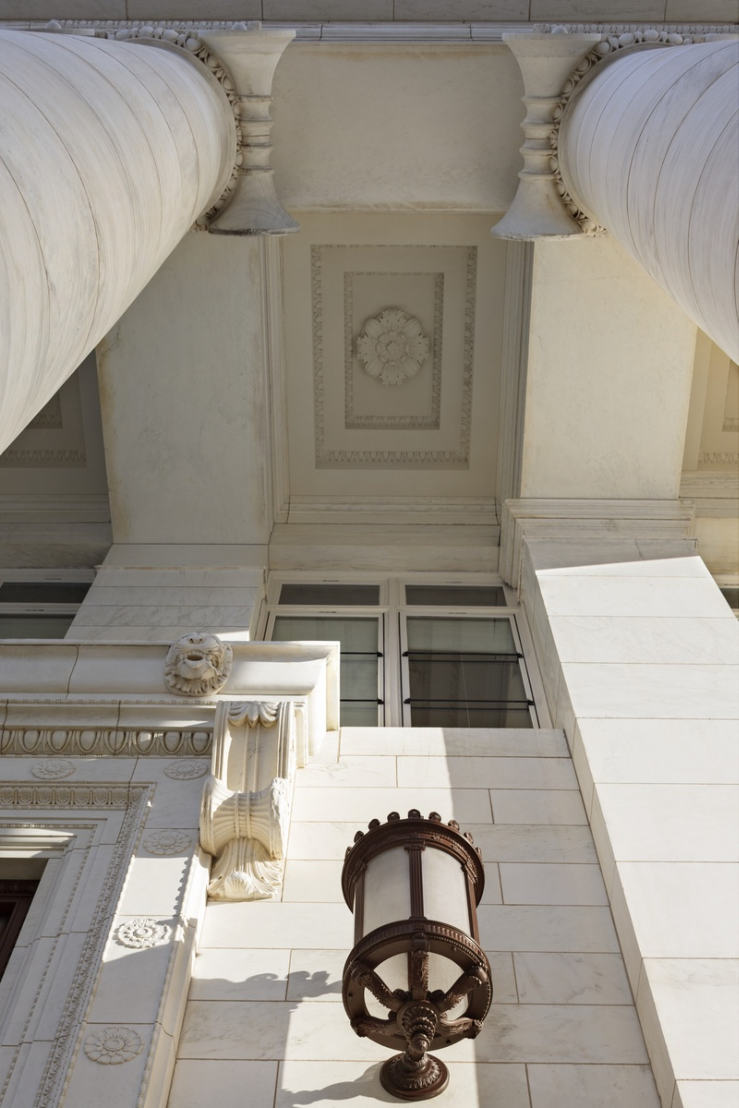 Exterior view of a building with classical architectural details, including large columns, ornate decorative elements, and a vintage street lamp mounted on the wall.