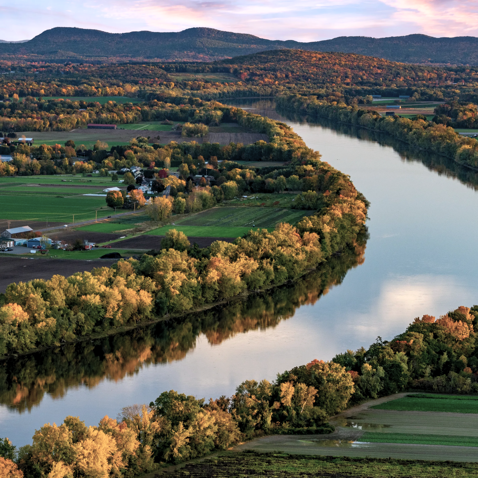 A scenic river view surrounded by autumn-colored trees, rolling hills, farm fields, and distant mountains under a partly cloudy sky.