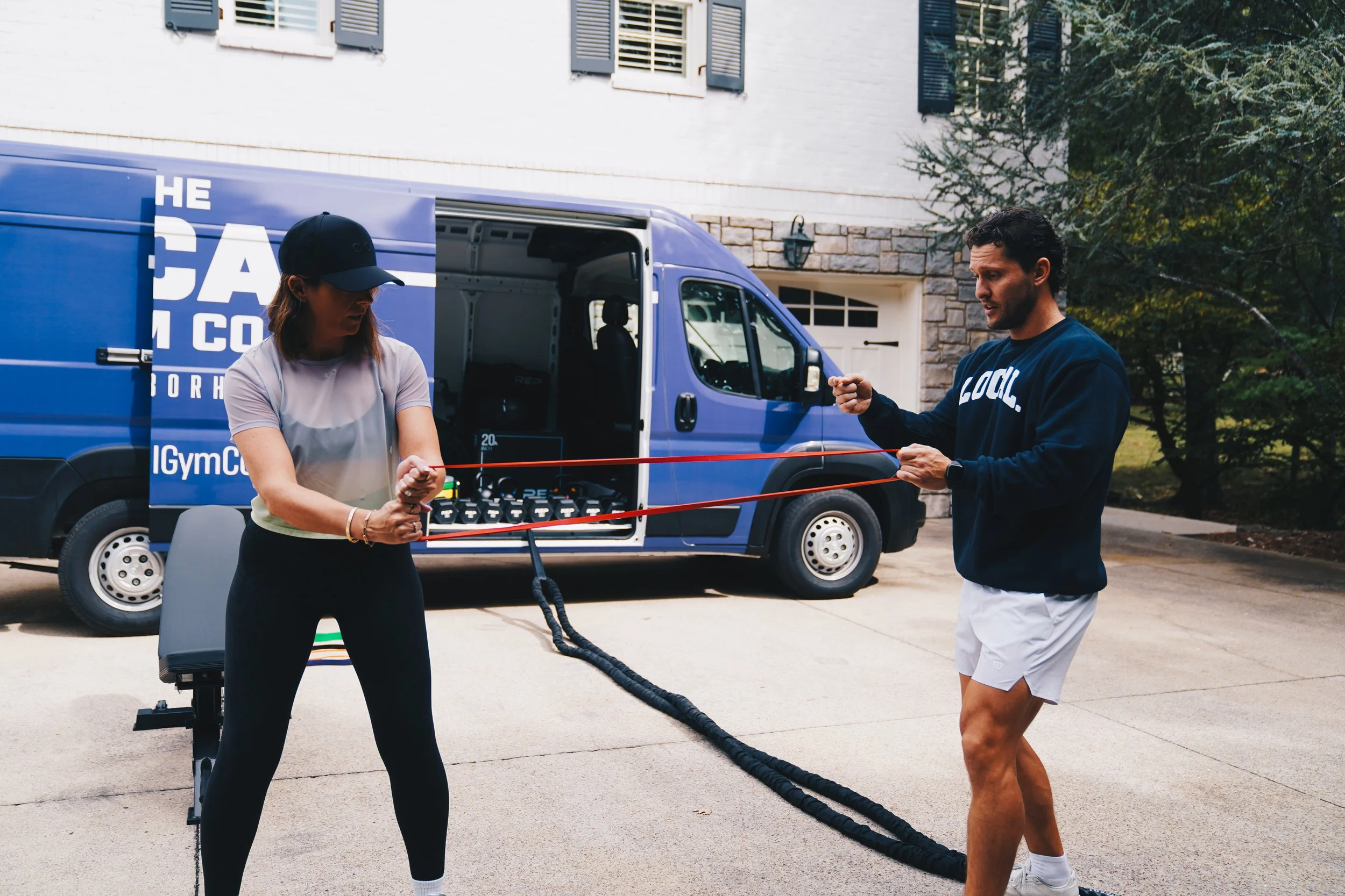A woman and a man are exercising outdoors in front of a blue delivery van. The woman is wearing a black cap and a light gray shirt, while the man is wearing a dark sweatshirt with the word 'LOCAL' on it and white shorts. They are holding a resistance band, practicing a workout together.