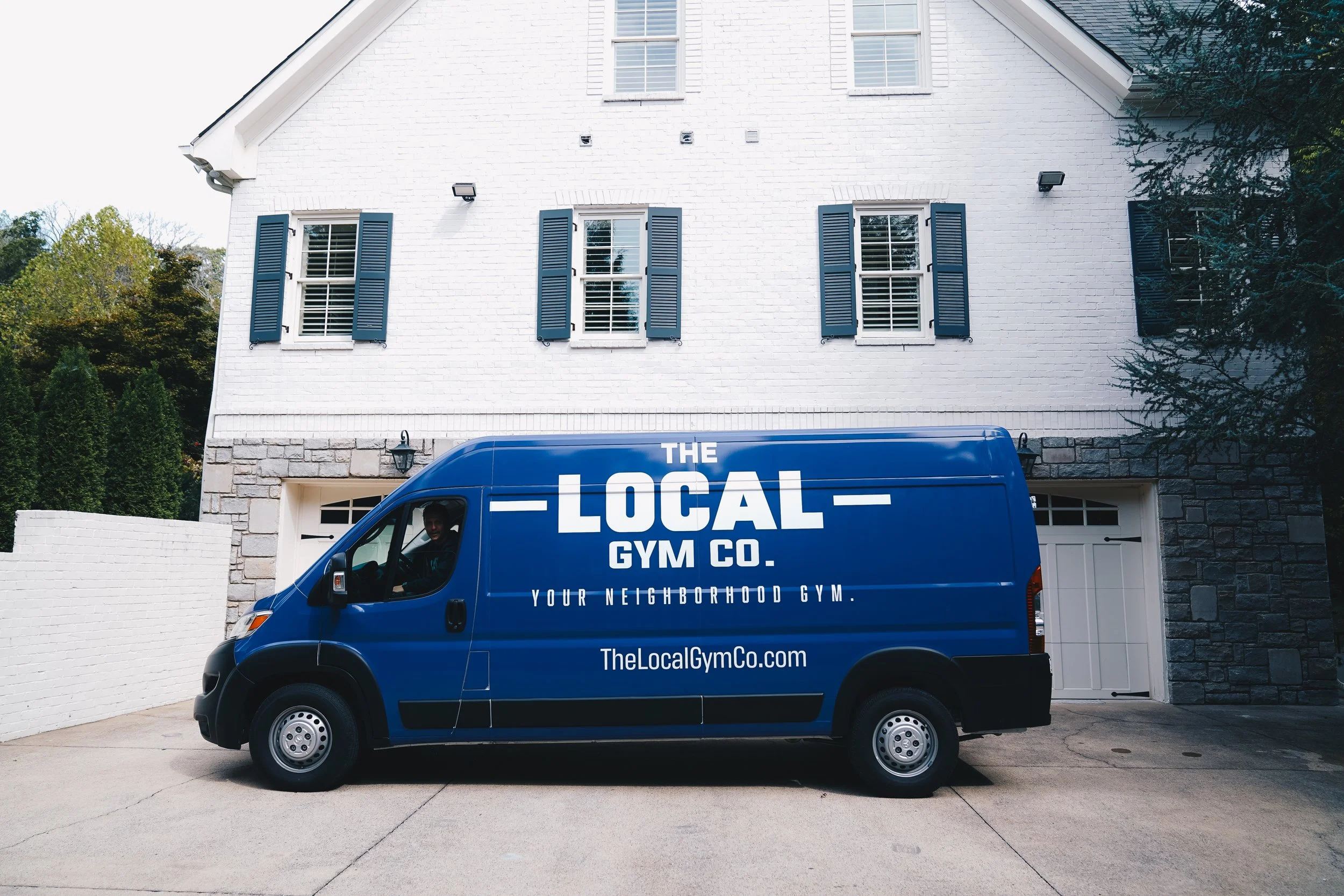 Blue delivery van parked in front of a white brick house with blue shutters and a stone base, advertising 'The Local Gym Co.' and the website 'TheLocalGymCo.com'.