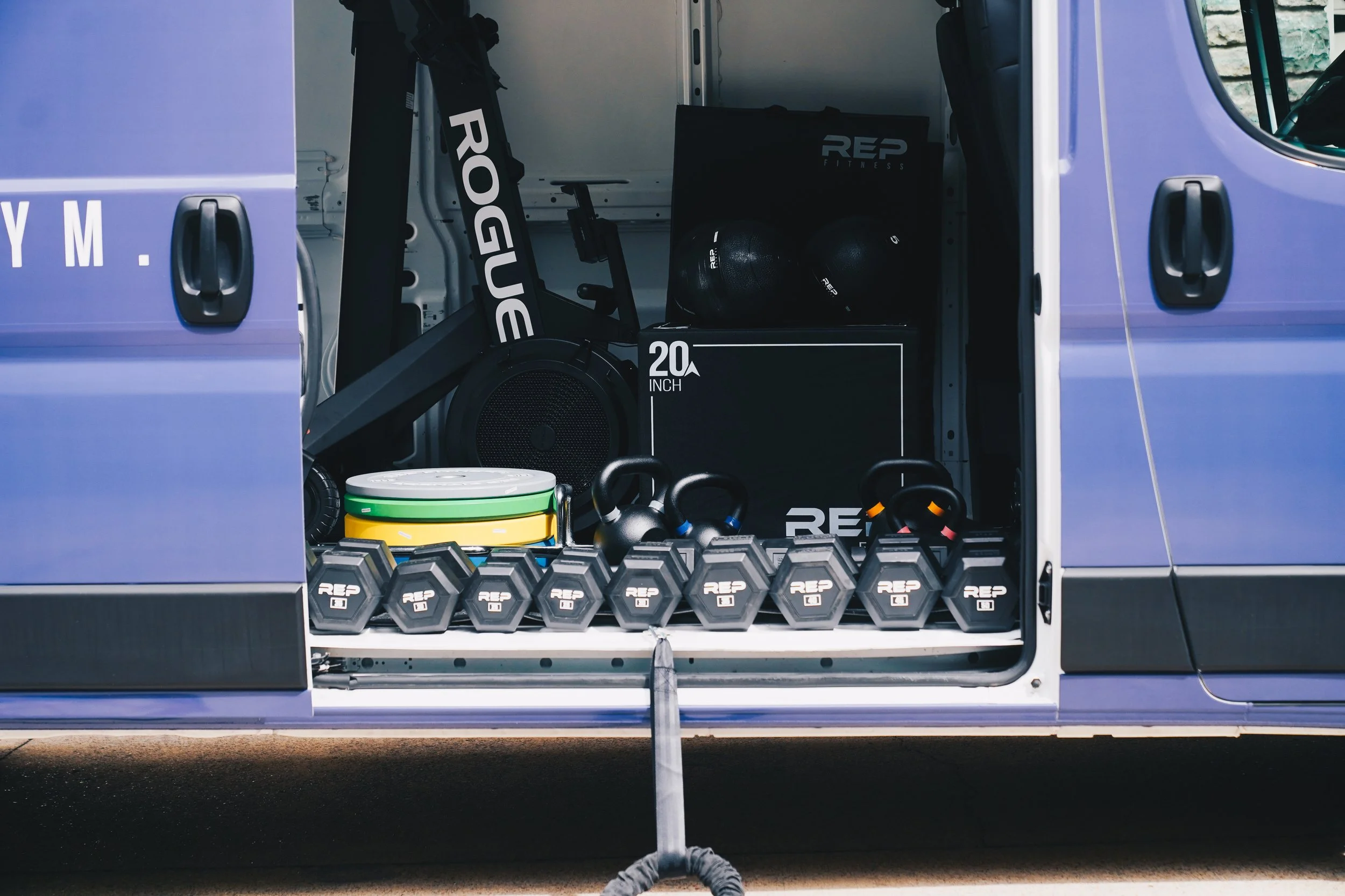 Open side of a van filled with fitness equipment including kettlebells, medicine balls, dumbbells, resistance bands, a foam roller, and a squat rack.
