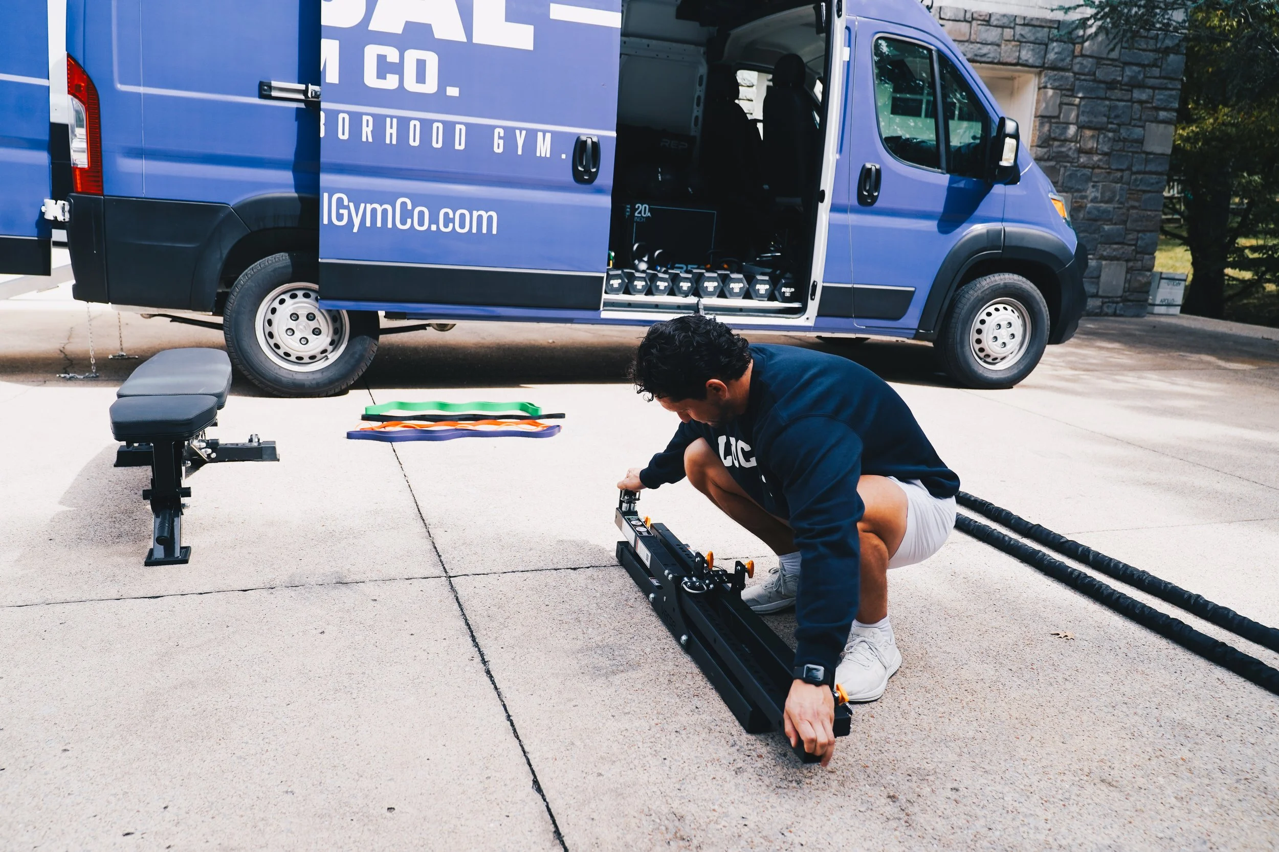 A man kneeling on the sidewalk adjusting equipment in front of a blue company van with the words 'Royal Norhood Gym' and the website 'IGymCo.com' printed on it. The van has its side door open, revealing workout equipment inside, including a row of kettlebells. There are exercise bands, a weight bench, and other fitness gear on the ground near him. A stone building and some trees are in the background.