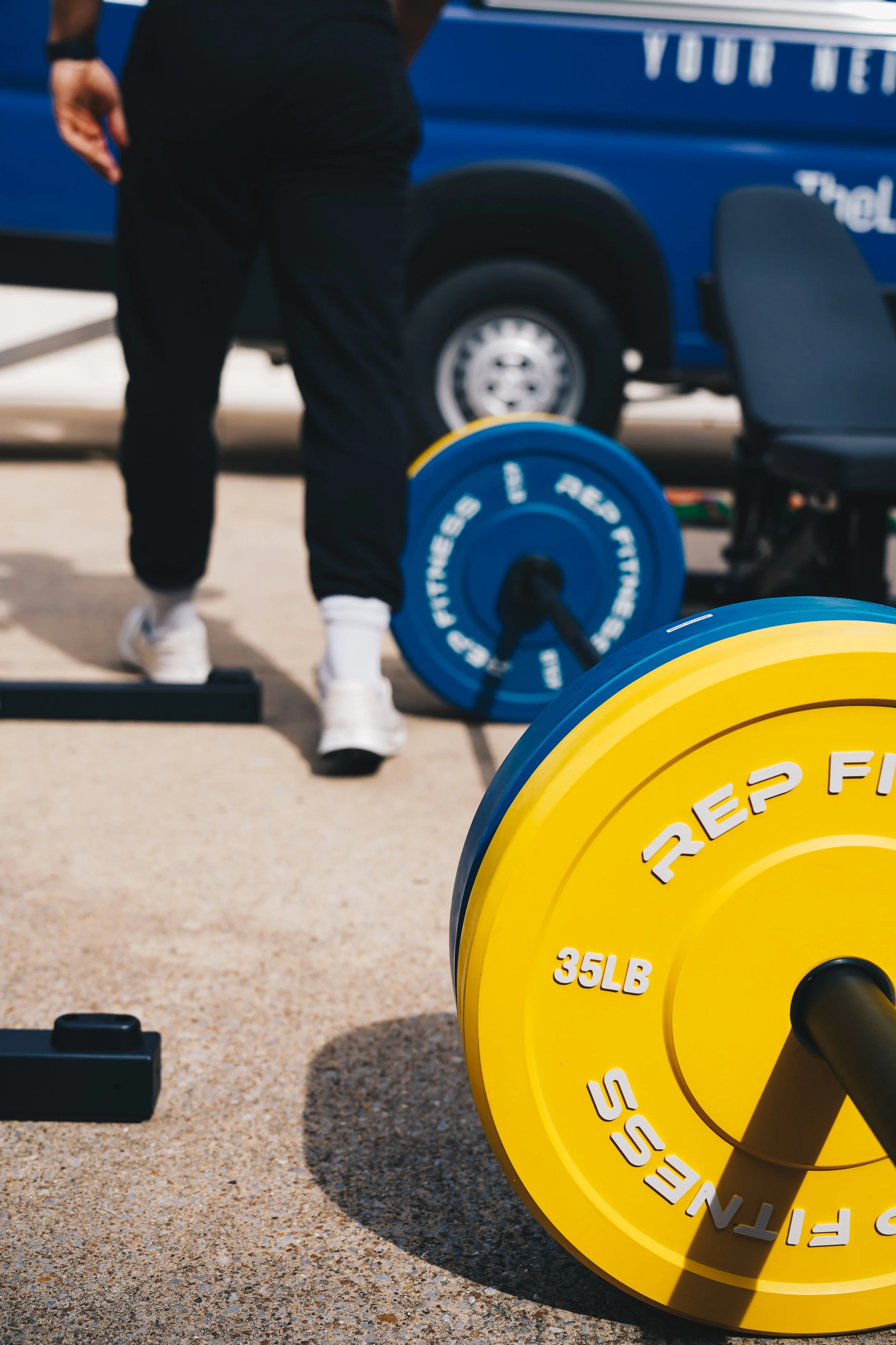 Close-up of yellow weight plate labeled 35LB on the ground during outdoor workout. In the background, a person in black pants and white sneakers is near blue and black gym equipment, with a blue vehicle partially visible behind.