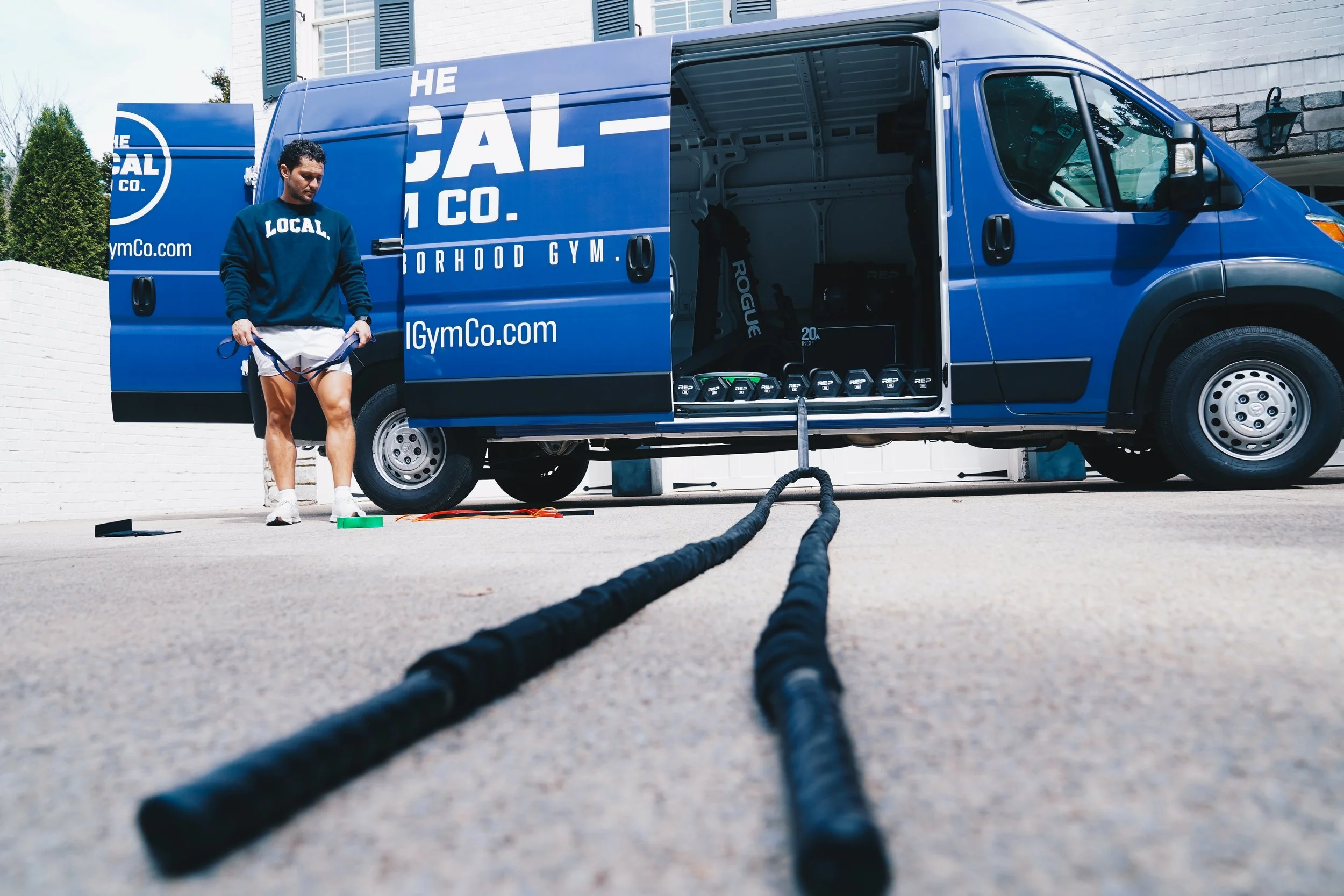 A man standing outside next to a blue delivery van with open side door, revealing gym equipment inside; the man holds a jump rope and wears a sweatshirt with 'LOCAL' printed on it.