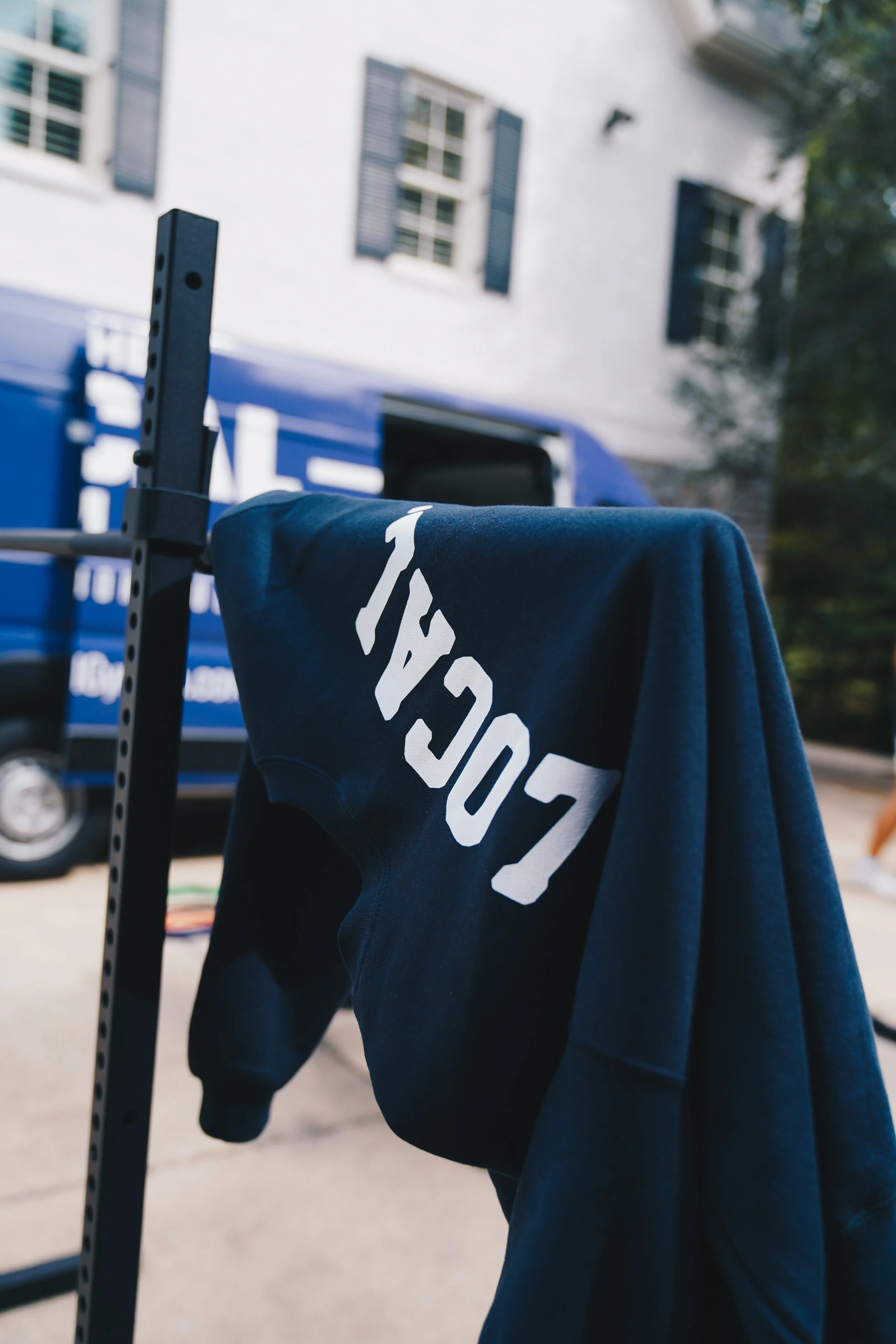 Close-up of a dark blue sweatshirt with white text hanging on a black metal rack, with a blurred background of a building with white walls and blue window shutters.