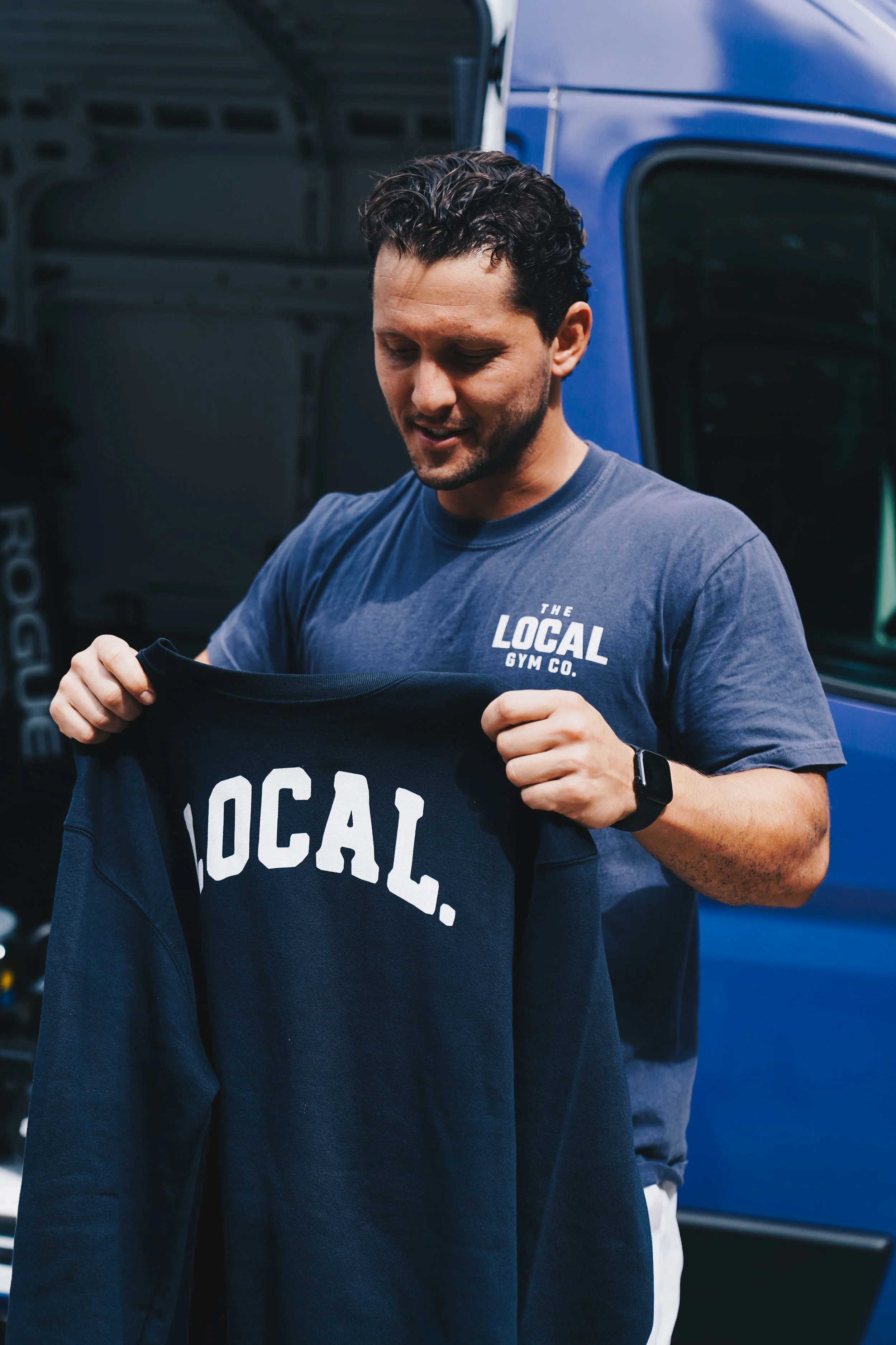A man with curly dark hair wearing a blue t-shirt that says "The Local Gym Co." is holding a black clothing item with white text that says "LOCAL." He is standing in front of a blue truck and appears to be looking down at the clothing item.