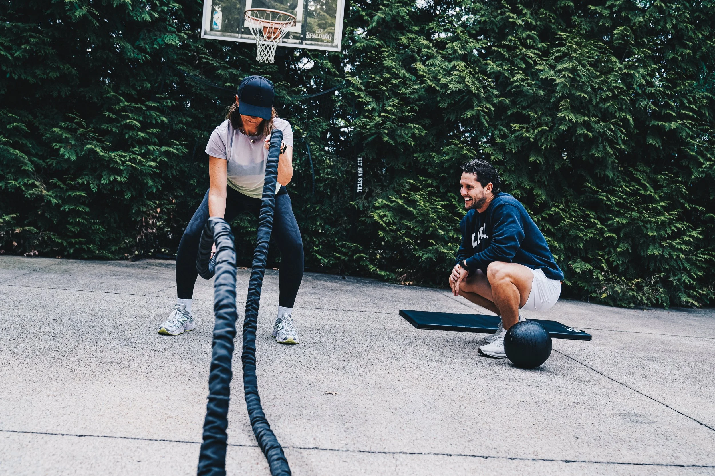 A woman in a cap and athletic clothes does battle ropes exercise while a man crouches nearby, smiling in a backyard with a basketball hoop and dense green trees.