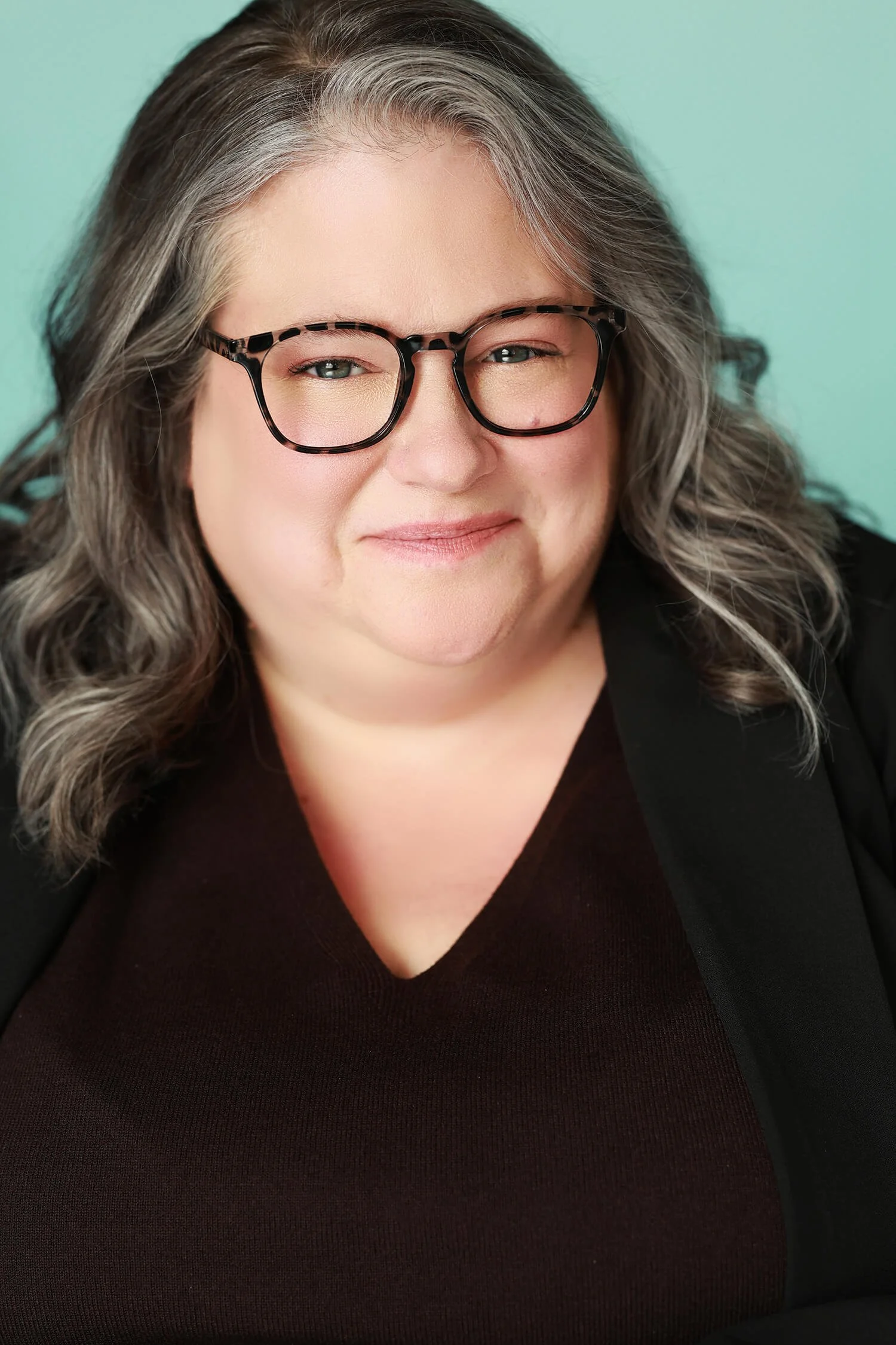 Close-up portrait of a woman with gray hair, wearing glasses, smiling, against a light mint-green background.
