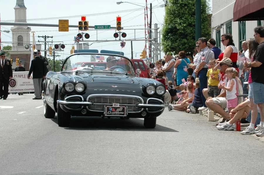 A vintage black Chevrolet Corvette convertible driving down a city street during a parade. Spectators, including children and adults, line the sidewalk watching the car pass. Traffic lights and street signs are visible above the parade, with a church steeple seen in the background.