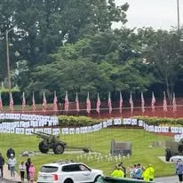 A park scene with American flags, trees, and a viewing area with chairs. Several cars are parked, and a group of people is gathered near a military vehicle.