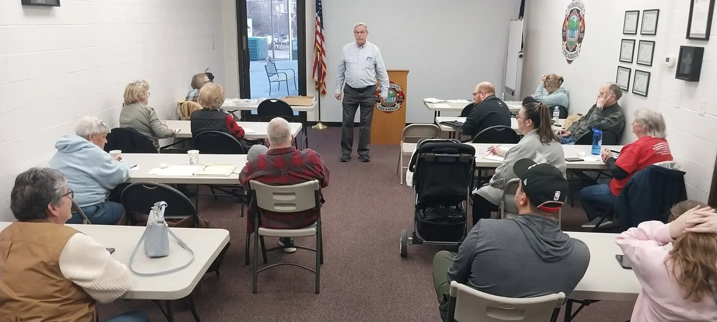 A man in a light shirt and dark pants stands at the front of a small meeting room, speaking to an audience of approximately 14 people seated at tables arranged in U-shape. The room has a white brick wall, framed documents, an American flag, and a door with a window showing a parking lot outside.