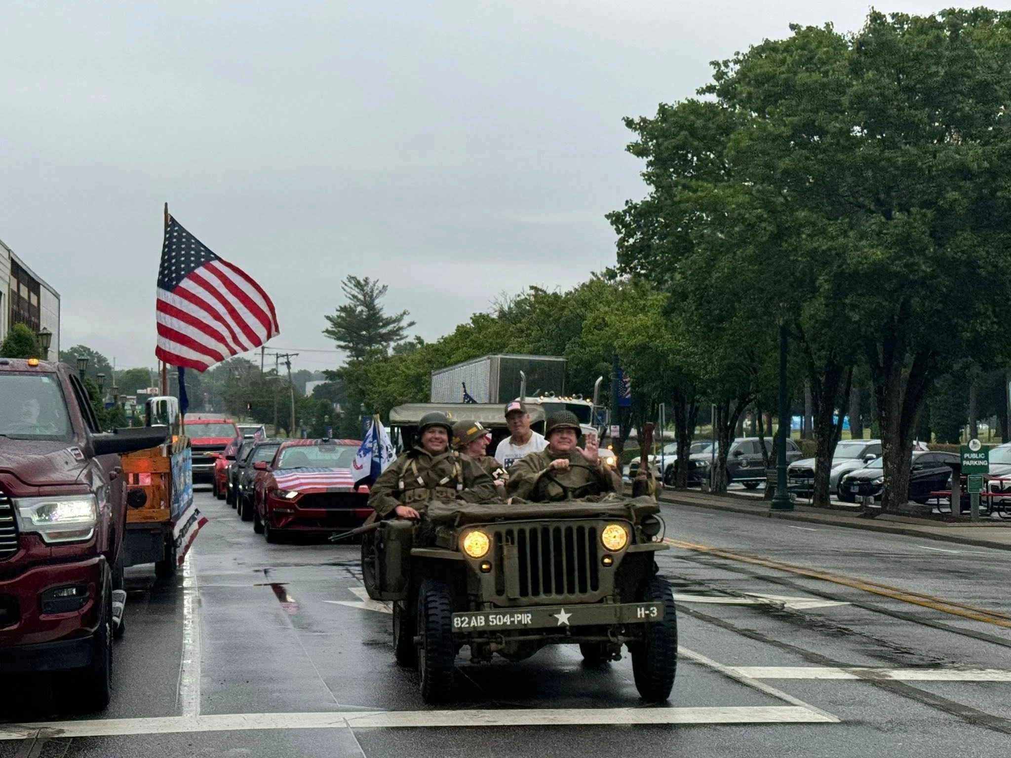 Veterans ride in a military jeep down a city street during a parade, with American flags and spectators along the sidewalk on a cloudy day.
