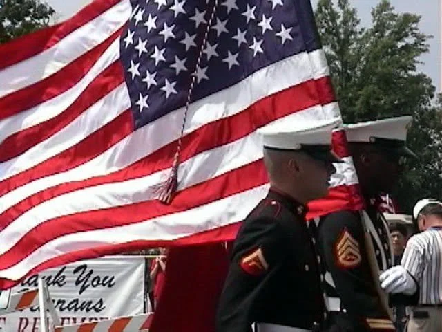 Two U.S. Marines in dress uniforms standing near a large American flag at a memorial event.