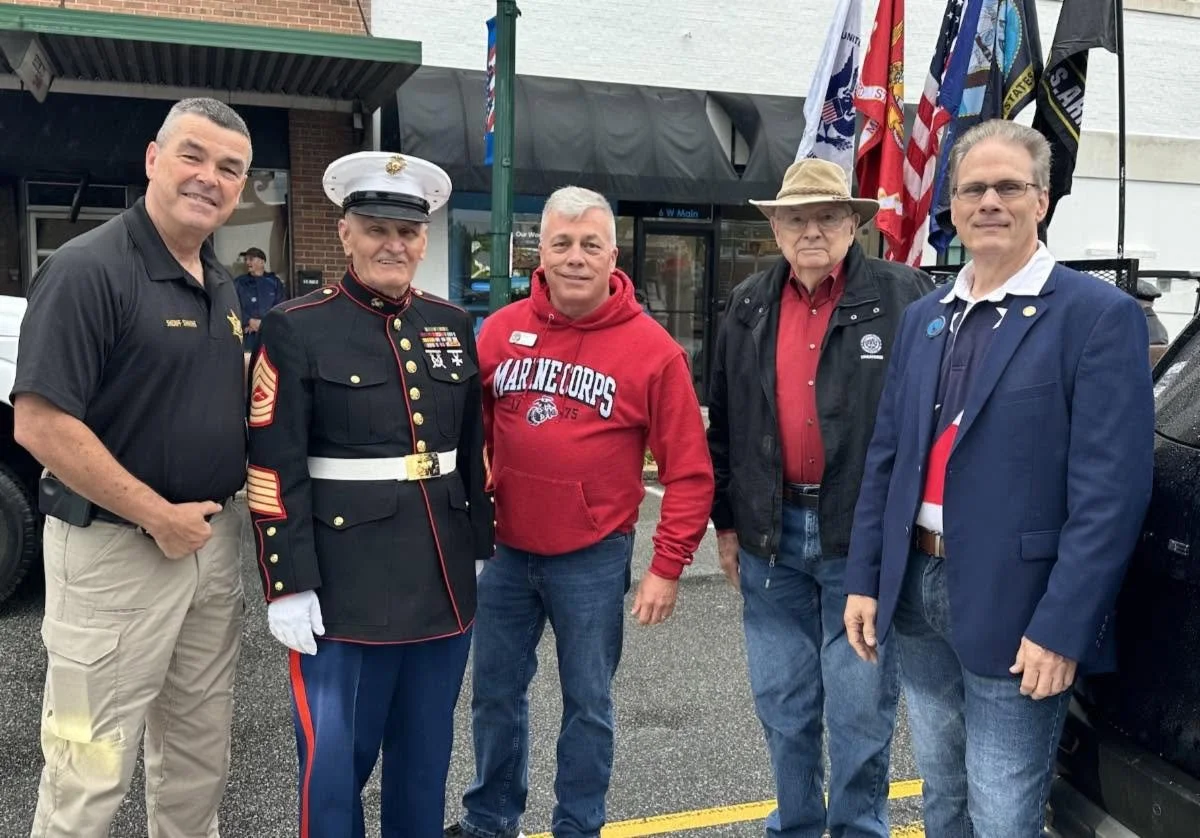Five men standing together outdoors at a patriotic event, with flags in the background. They include a uniformed Marine and men in casual and semi-formal attire.