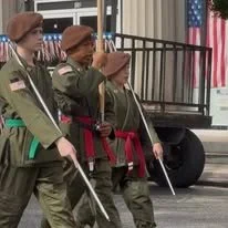 Four soldiers in uniform marching, carrying flags and rifles during a parade.