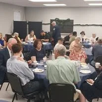 A group of people sitting at round tables in a conference room, listening to a speaker standing at the front near a blackboard or screen.