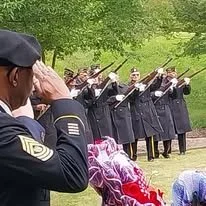 Military band performing outdoors with musicians in uniform playing instruments, and a person in the foreground taking a photo or video.