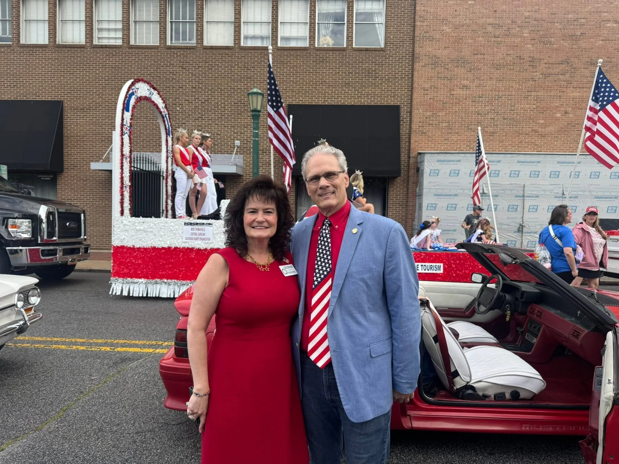 A man and woman dressed in red, white, and blue patriotic clothing standing in front of a parade float decorated with American flags and patriotic colors. The float is decorated with a large white star, red and white flowers, and has three women in red, white, and blue outfits standing on it. The background shows a street scene with parked cars, onlookers, and a brick building.