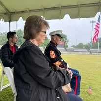 Three people sitting under a white tent, with a man in a uniform and a woman in a black jacket, gazing toward a field.