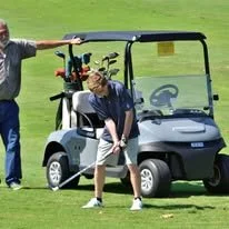 Two boys on a golf course, one standing and the other preparing to hit a golf ball, with a golf cart in the background.