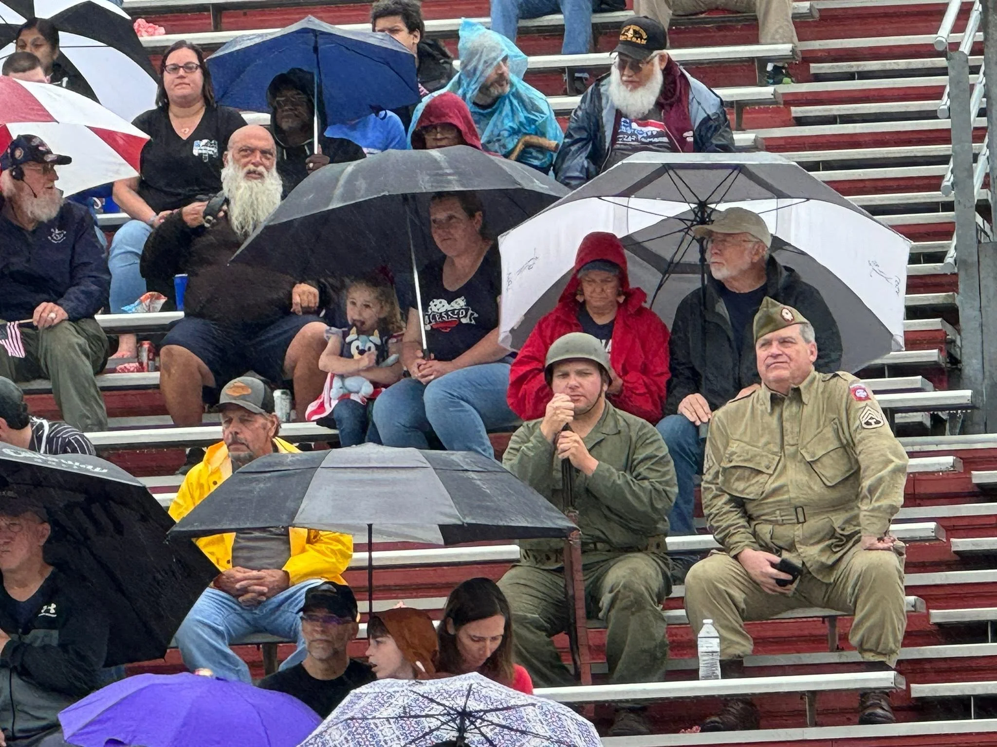 People sitting on bleachers holding umbrellas on a rainy day, including veterans and civilians, some wearing military uniforms and hats.