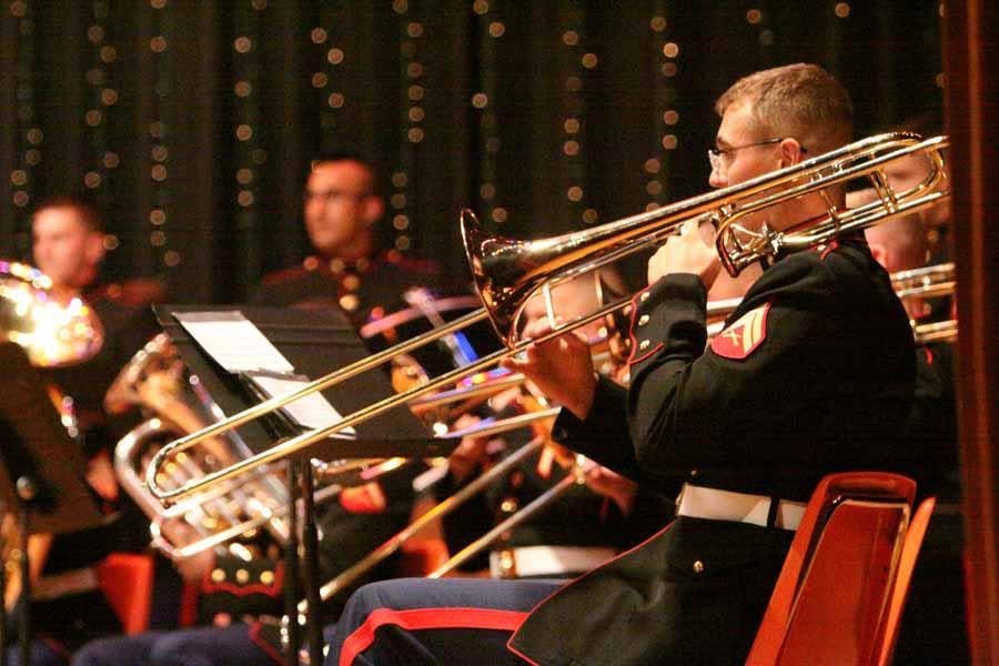 A young man in military uniform playing a trombone during a band performance, with other band members visible in the background on a stage with black curtains decorated with small lights.