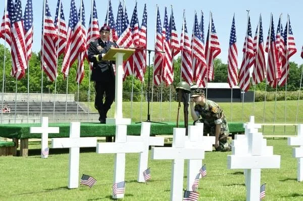 A memorial ceremony at a cemetery with U.S. flags in the background. One person in a military uniform is reading at a podium, while another is kneeling next to a grave, placing a small American flag on a white cross.