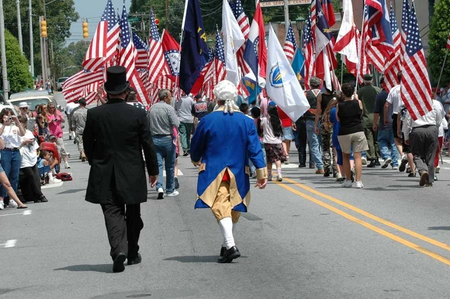 People participating in a parade with American flags, including a person dressed as George Washington, and a crowd of spectators watching on the sides.